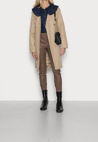 Trench coat beige con colletto arricciato in denim blu navy, abbinato a pantaloni di pelle marrone e stivaletti neri. Una piccola borsa nera completa il look.