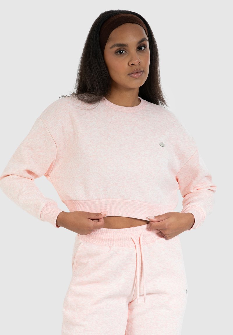 Felpa corta rosa chiaro con un motivo testurizzato, polsini a costine e un piccolo logo. Abbinata a pantaloni joggers rosa chiaro coordinati.