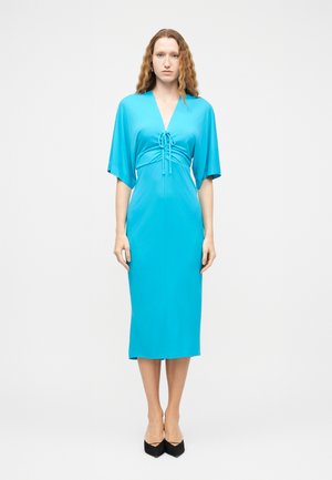 Femme aux longs cheveux ondulés, debout de face, portant une robe midi bleu vif à décolleté en V et des chaussures noires à bout pointu.