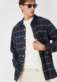 Camisa de franela a cuadros en tonos azul marino con patrones de cuadros blancos y rojos, con botones en la parte delantera, cuello tipo polo y mangas largas, llevada sobre un suéter ligero.
