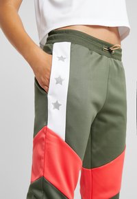 Olivgrön atletisk jogger med orange och vita paneler. Har silverstjärnor som dekorationer på en sidoficka, elastisk midja och dragsko.