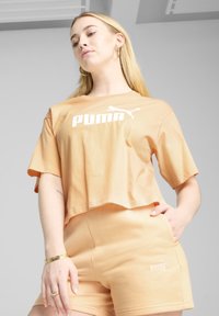 Lys ferskenfarvet crop top med hvid "PUMA" logo, parret med matchende shorts med et lille logo. Blødt stof, afslappet pasform. Guld smykkeaccent.