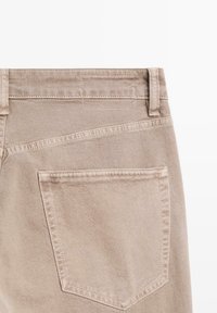 Beżowe dżinsy z średnim stanem, z pojedynczą kieszenią z tyłu i widocznymi detalami szwów. Gładka tekstura z klasycznym wzornictwem.