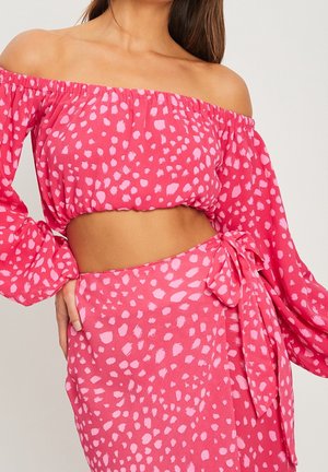 Frau trägt ein rosa schulterfreies Crop-Top und einen passenden Wickelrock mit unregelmäßigem hellrosa Punktemuster.