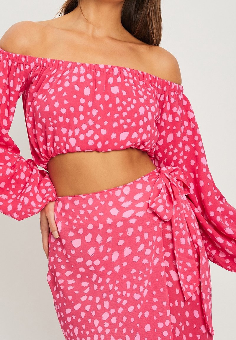 Femme portant un crop top rose à épaules dénudées et une jupe portefeuille assortie avec un motif de points irréguliers rose clair.