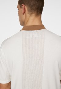Polo bianco con colletto marrone, dotato di un pannello posteriore testurizzato e traspirante con piccole perforazioni. Design semplice e sportivo.