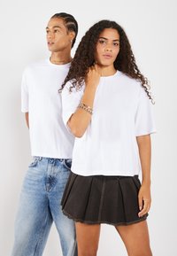 Białe t-shirty z krótkim rękawem noszone przez dwie osoby, w parze z ciemną plisowaną spódnicą i jasnoniebieskimi dżinsami. Prosty, casualowy design.