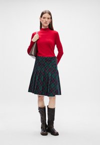 Κόκκινο, μακρυμάνικο, εφαρμοστό πουλόβερ με λαιμόκοψη, συνδυασμένο με πράσινη καρό, πλισέ φούστα και καφέ δερμάτινες μπότες μέχρι το γόνατο.