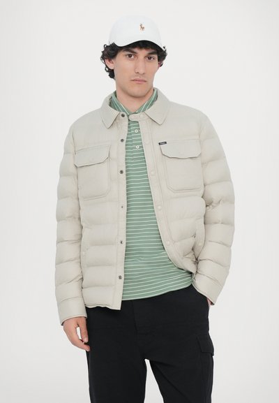 Lys beige dunjakke med trykknapper, to brystlommer, båret over en grøn og hvid stribet polo-shirt, kombineret med sorte bukser. Hvid kasket.