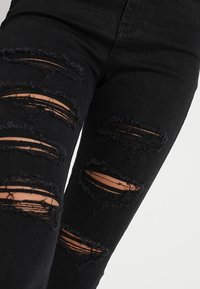 Jeans de mezclilla negros desgastados con múltiples grandes desgarros que revelan la piel debajo. La textura parece deshilachada a lo largo de los bordes de los desgarros.