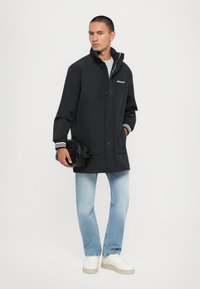 Black lightweight jacket with a high collar, snap buttons, and striped cuffs. Paired with light blue jeans and white sneakers.