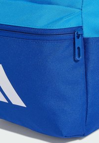 Modrý látkový batoh s texturovaným povrchem, zipovou kapsou a bílými логy na bočním panelu.