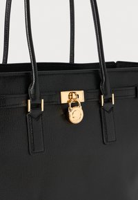 Bolso de cuero negro con dos asas y un detalle de candado circular dorado en el broche central delantero.