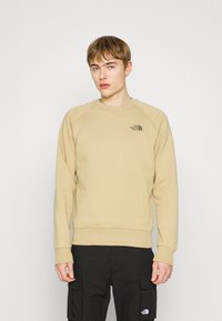 Sudadera beige con largas mangas raglán, puños y cuello acanalados. Presenta un pequeño logo negro en el lado izquierdo del pecho. La tela parece suave.