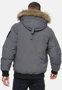 Graue Pufferjacke mit einem Kunstpelzbesatz an der Kapuze, gerippten schwarzen Bündchen und Saum sowie einem Patch an den linken Ärmel. Verfügt über ein flaches Nahtdesign.