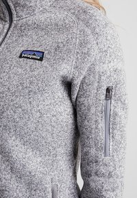 Grå fleecejacka med hög krage, dragkedjeficka på ärmen och en Patagonia-logopatch på bröstet. Mjuk textur och stickad mönster.