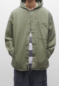 Chaqueta verde oliva con capucha, cierre de botones y un bolsillo en el pecho. Hecha de tela suave, la lleva puesta sobre una camisa a rayas en gris y blanco.