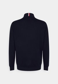 Granatowy sweter z wysokim kołnierzem, posiada czerwono-białą paski przy kołnierzu, długie rękawy, ściągacze na mankietach i dole, miękka dzianinowa struktura.