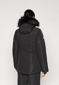 Μαύρο μονωμένο μπουφάν με επένδυση τεχνητής γούνας στον γιακά, ομαλή υφή, κουκούλα και επιμηκυμένο πίσω τελείωμα· λογότυπο στο μανίκι, μινιμαλιστικός σχεδιασμός.