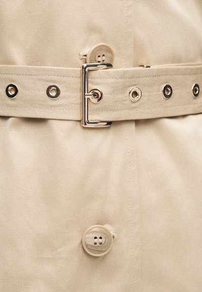 Ceinture en tissu beige avec boucle en métal et œillets, attachée autour d'un manteau beige assorti avec deux grands boutons.