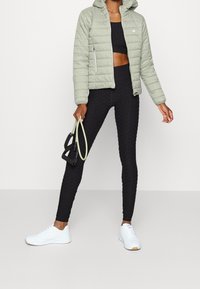Femme portant une veste matelassée vert sauge, un ensemble de sport noir texturé, des baskets blanches, tenant une bande de résistance devant un fond uni.