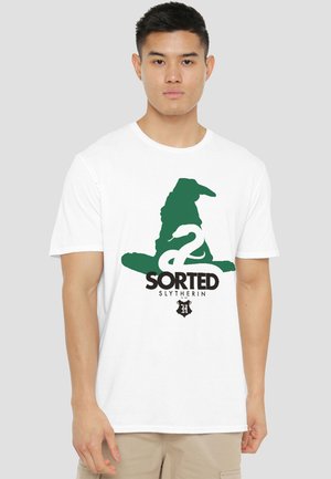 Young man wearing a white T-shirt with a green sorting hat, a white snake, and text reading "SORTED SLYTHERIN" on the front.