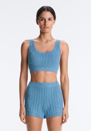 Femeie purtând un top scurt tricotat bleu deschis fără mâneci și pantaloni scurți în talie înaltă asortați, stând în fața unui fundal alb.
