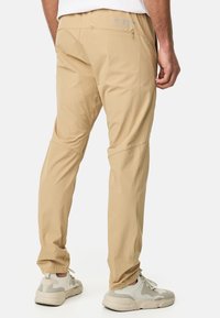 Pantalons légers beiges avec une texture lisse, dotés de poches latérales et d'une coupe décontractée, associés à des baskets grises et blanches.