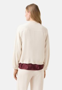 Kremowa bomberka z elastycznymi mankietami, z satynowym bordowym dołem. Gładka faktura, luźny krój i subtelny detal w postaci szwu na plecach.