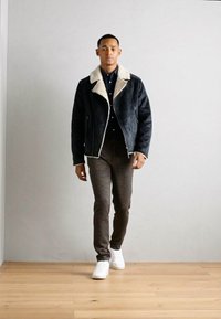 Black shearling jacket with white collar, navy button-up shirt, brown patterned pants, and white sneakers, standing against a light wall.