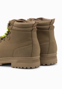 Botas alta de color tostado con una textura suave, suelas de goma gruesa y cordones de neón verde. Incluyen un cuello acolchado y detalles de costura reforzada.