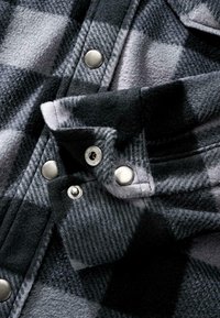 Gros plan d'un tissu à carreaux noir et gris avec des boutons-pression métalliques argentés sur une manche de vêtement et la couture avant.