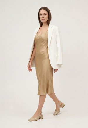 Vrouw loopt in een gouden satijnen slipjurk, beige hak slingback schoenen en een wit blazer over haar schouders gedrapeerd.