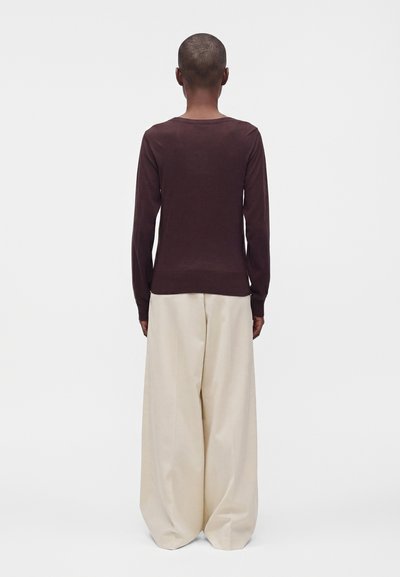 Personne aux cheveux courts debout, dos à la caméra, portant un haut à manches longues marron foncé et un pantalon large beige sur un fond uni.