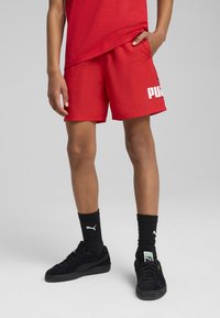 Pantalones cortos deportivos rojos con el logo blanco de "PUMA", combinados con zapatillas negras y calcetas negras de canalé. Tejido suave, corte holgado, longitud por encima de la rodilla.