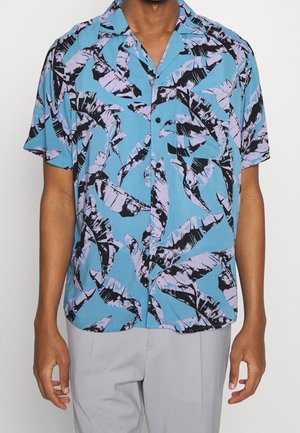 Homme portant une chemise à manches courtes avec un fond bleu et des motifs de feuilles abstraites noires et violet clair.