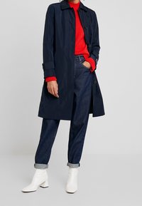 Granatowy trencz z kołnierzem, zapięciem na guziki i długim rękawem, zestawiony z czerwonym swetrem dzianinowym, niebieskimi dżinsami i białymi botkami za kostkę.