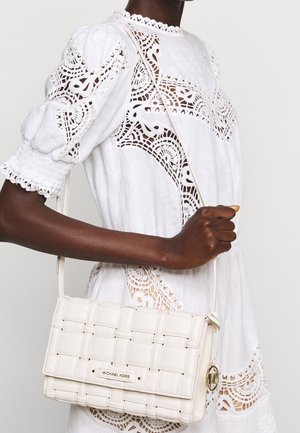 Vrouw in een witte kanten jurk die een wit gevlochten leren Michael Kors crossbodytasje met gouden logo hanger vasthoudt.