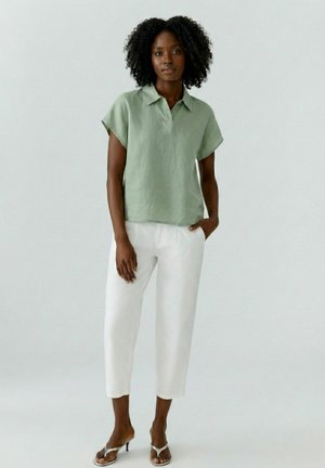 Femme aux cheveux naturellement bouclés portant une chemise vert clair à manches courtes, un pantalon blanc court et des sandales argentées à talons, debout devant un fond uni.