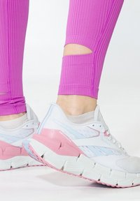 Zapatillas Reebok blancas y rosas con suela texturizada, que presentan un diseño robusto y detalles en azul claro, combinadas con leggings rosa acanalados.