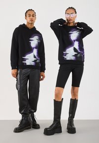 Svart hettegenser med et lilla abstrakt design, sammen med svarte skinnbukser og teksturerte svarte støvler, båret av to modeller.