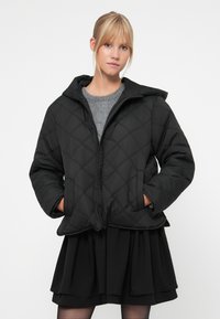 Črna prešita jakna s kapuco, ki ima diagonalni šiv, z zadrgo spredaj in stranskimi žepi. Oblečena čez sivo majico in črno krilo.