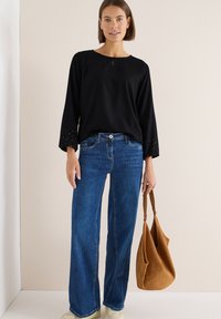 Schwarze Bluse mit einem Schlüssellochdetail und verzierten Ärmeln, kombiniert mit blauen weit geschnittenen Jeans und einer braunen, lässigen Handtasche.