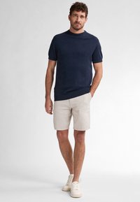 Maglione a maniche corte blu navy abbinato a pantaloni corti a righe beige. Il modello indossa sneakers bianche e si trova di fronte a uno sfondo neutro.