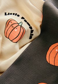 Twee gelaagde stoffen stukken: de ene crème met een glimlachende pompoenafbeelding en de tekst "Little Pumpkin," de andere donkergrijs met herhalende pompoenpatronen.