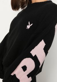 Czarny sweter z różowym logo królika Playboy na klatce piersiowej i dużymi różowymi literami na rękawie, noszony przez osobę z kolczykiem w kształcie koła.