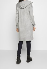 Osoba ubrana w długi szary kardigan z kapturem, ciemne skinny pants i czarne botki za kostkę, stojąca na tle jasnej, jednolitej powierzchni.