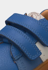 Chaussure enfant en cuir bleu avec deux bandes Velcro et coutures visibles, semelle beige clair, et étiquette beige avec le nom de la marque sur la languette.