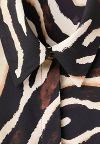 Črno-kremna srajca z ovratnikom, ki ima abstraktni živalski tisk z rjavimi poudarki. Gladek material z rahlo ohlapnim krojem in zapiranjem na gumbe.