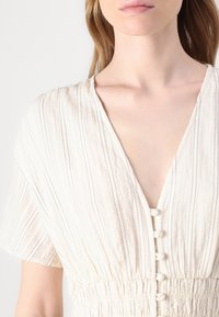 Abito beige chiaro realizzato in tessuto texturizzato con strisce verticali. Presenta una scollatura a V e bottoni sul davanti. Vita elasticizzata.